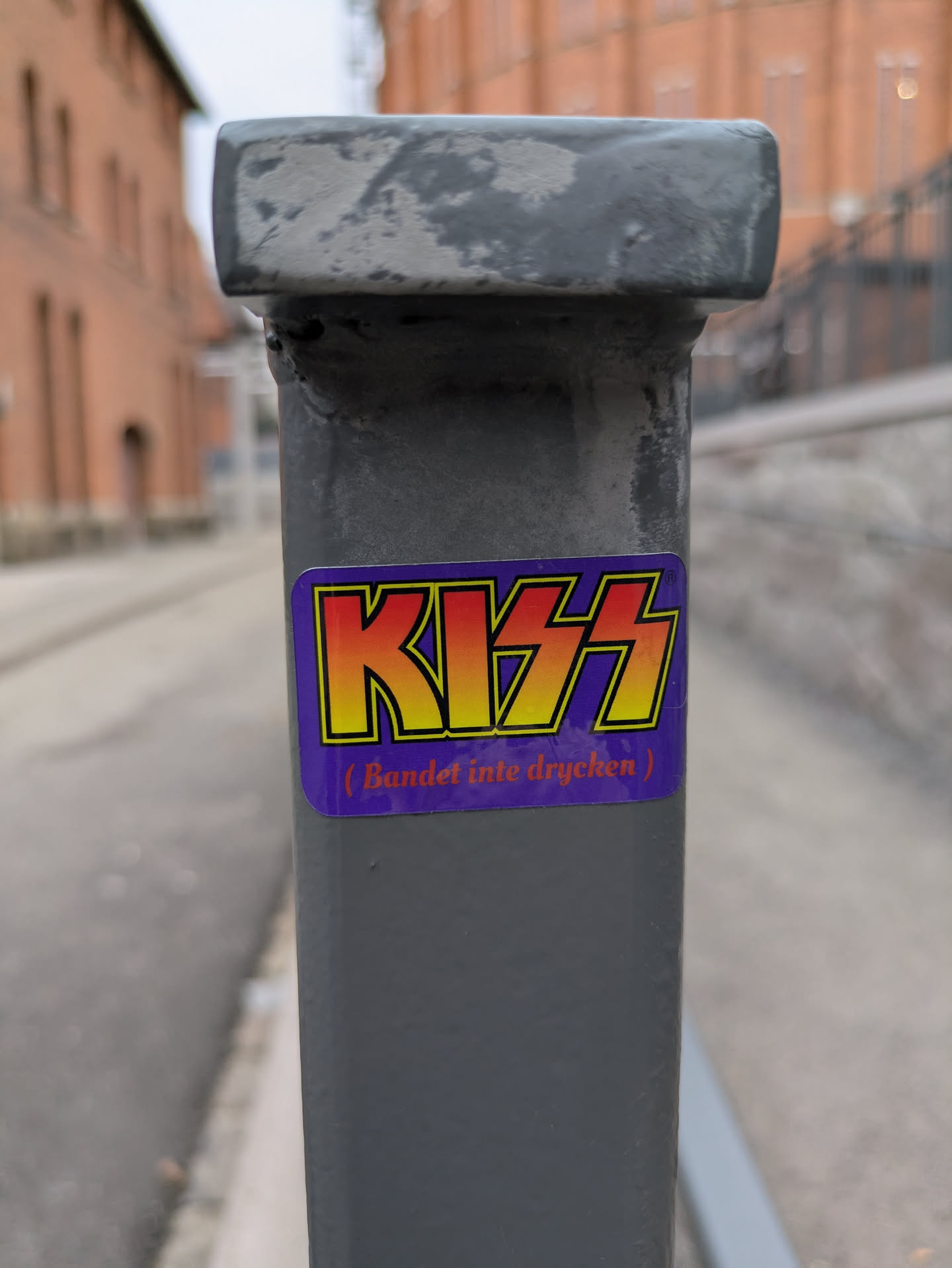 A sticker featuring the iconic KISS logo is affixed to a gray metal pole. Below the logo, smaller text reads '(Bandet inte drycken)', which translates to '(The band, not the drink)' in Swedish.