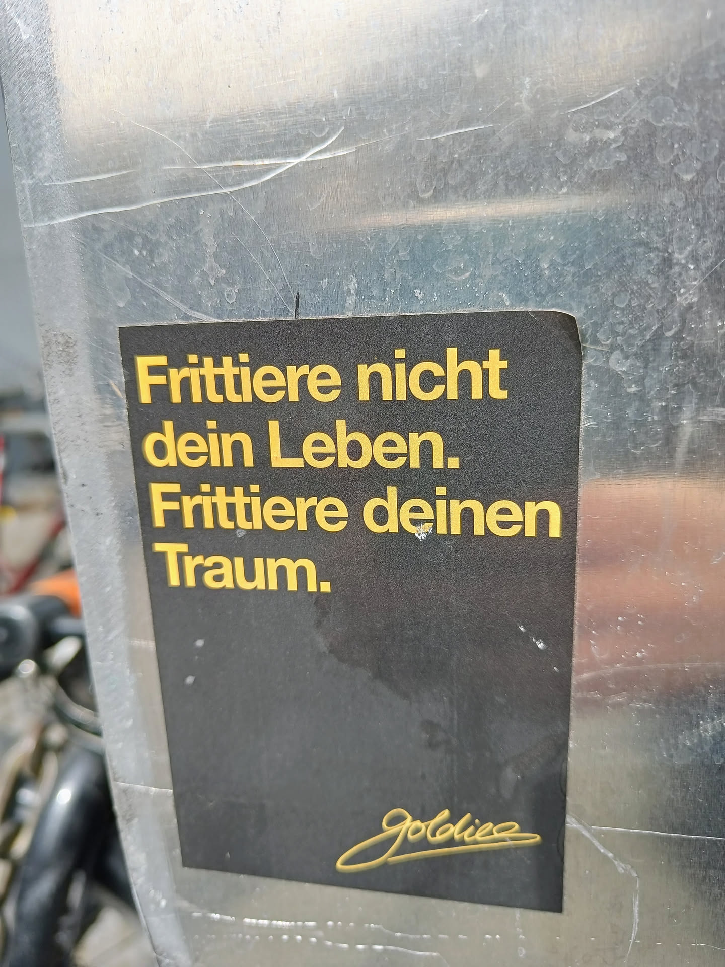 Black and gold sticker with a motivational quote in German. The text translates to "Don't fry your life. Fry your dream."  It's signed with a stylized signature, possibly the artist's name.
