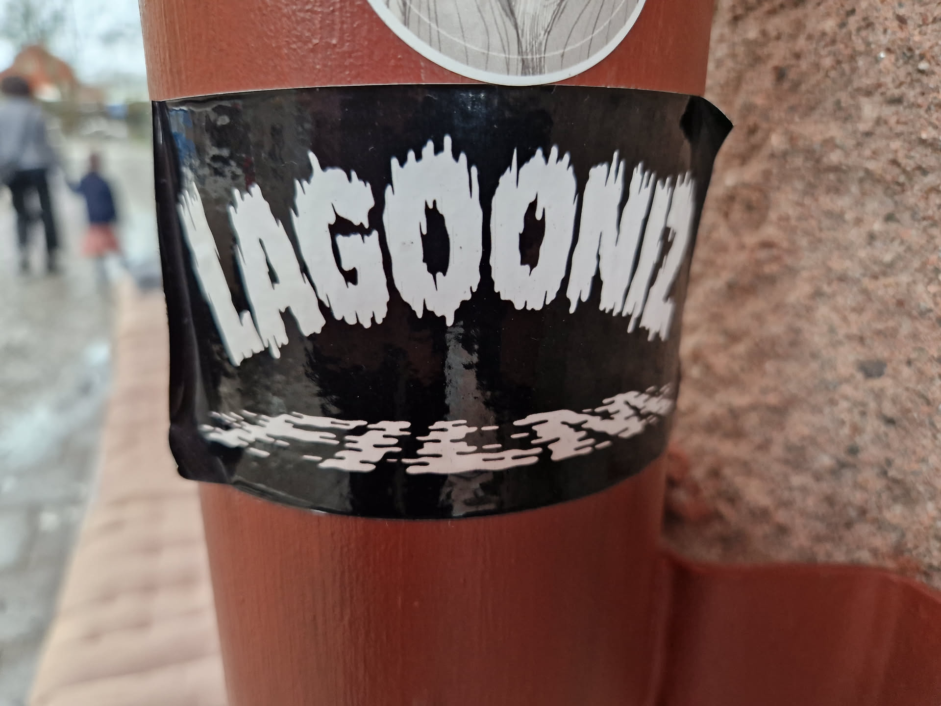 A black and white sticker with the word "LAGOONIZ" in stylized, dripping text.  There is a smaller graphic at the bottom. It's stuck to a reddish-brown pole.