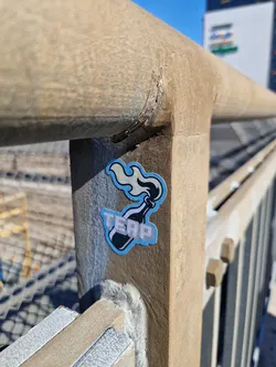 A small sticker depicting a Molotov cocktail with the word 'TERP' printed on it, affixed to a metal railing. The sticker is light blue and white with a pixelated style. The background shows a metal railing and a blurry industrial setting.
