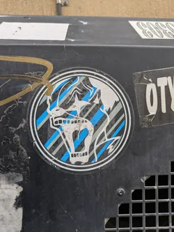 A round sticker featuring a skull with a screaming expression. The skull is depicted in white against a background of blue and gray diagonal stripes within a dark circular border. The style is reminiscent of graphic design from the 80s or 90s.