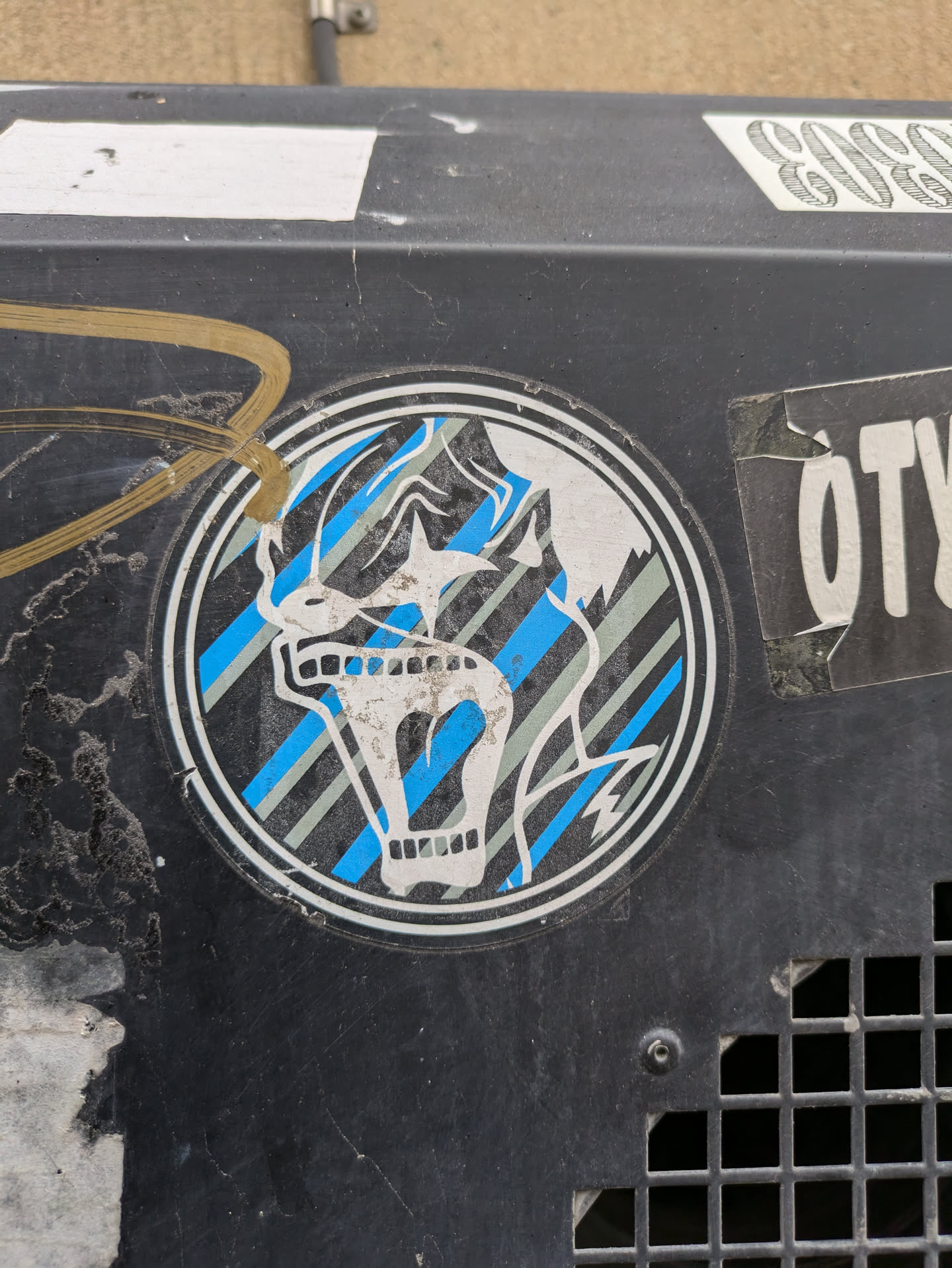 A round sticker featuring a skull with a screaming expression. The skull is depicted in white against a background of blue and gray diagonal stripes within a dark circular border. The style is reminiscent of graphic design from the 80s or 90s.