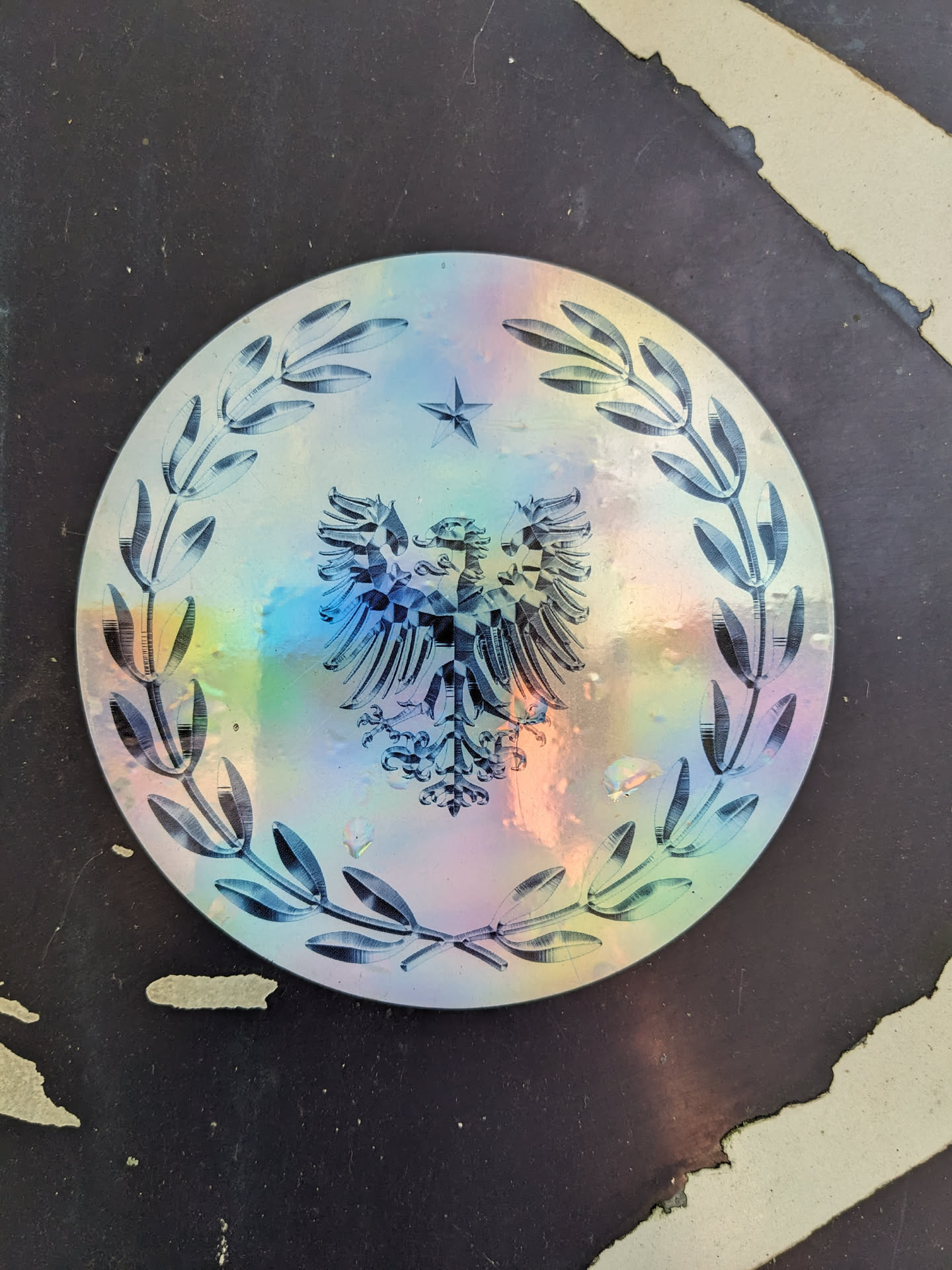 This eye-catching holographic sticker showcases an intricate design. A double-headed eagle, symbolic of power and authority, takes center stage, surrounded by olive branches representing peace and victory. The holographic effect creates a mesmerizing iridescent shimmer.