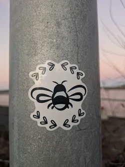 A cute bee sticker with a heart border. The bee is black and white and the hearts are a light gray. The sticker is on a metal pole.