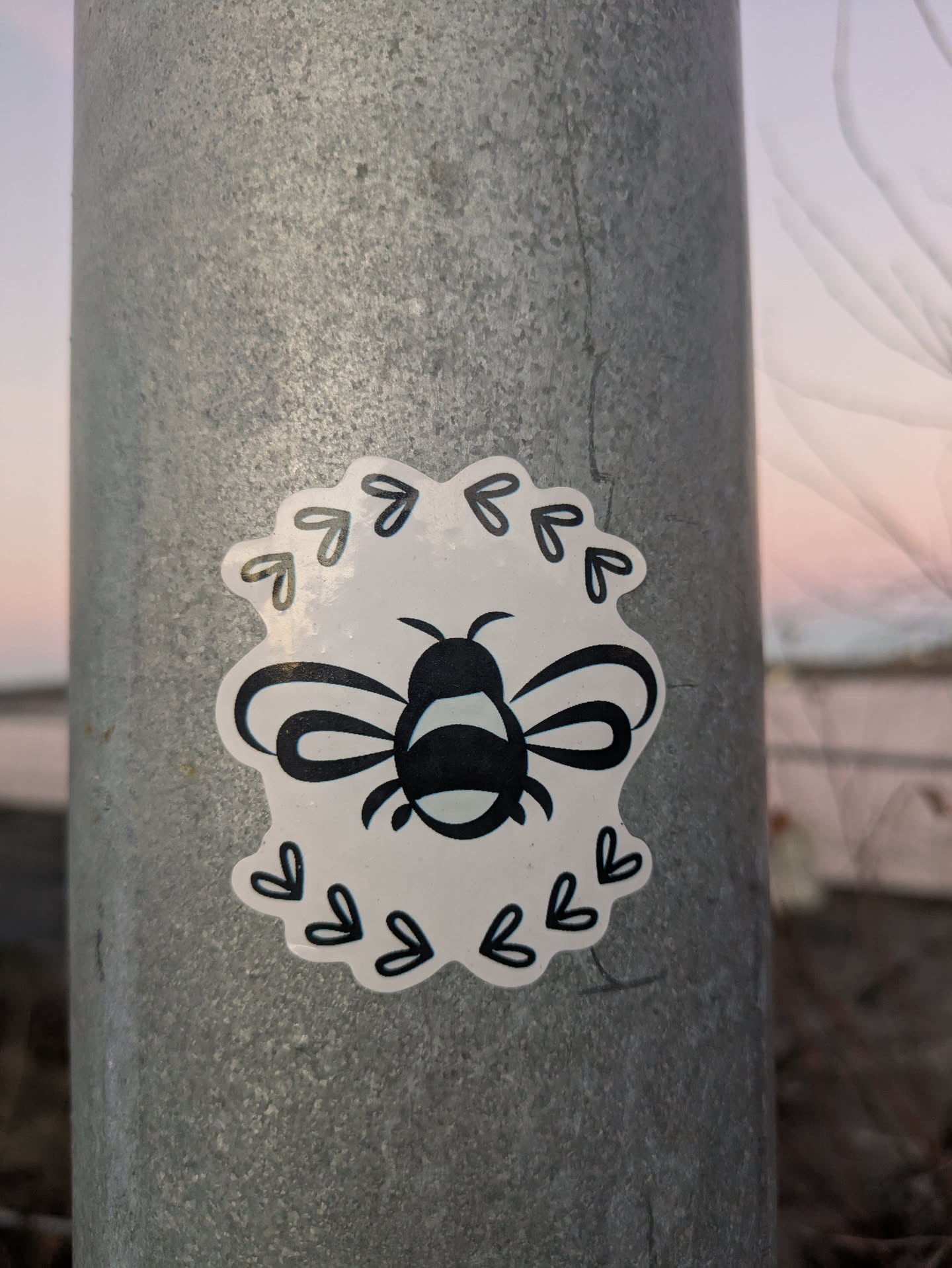 A cute bee sticker with a heart border. The bee is black and white and the hearts are a light gray. The sticker is on a metal pole.