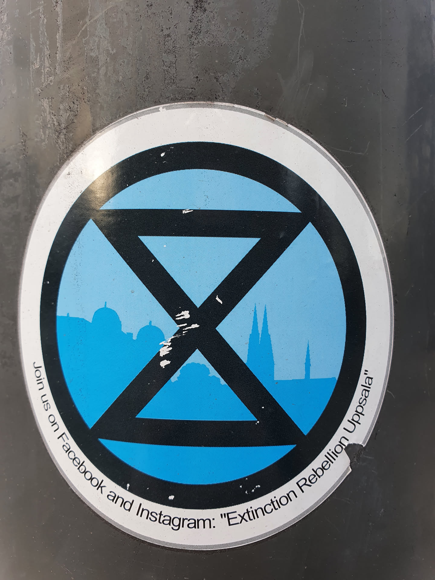 A sticker featuring the Extinction Rebellion symbol, a stylized hourglass, with a cityscape in the background.  The sticker also includes a call to join on Facebook and Instagram.