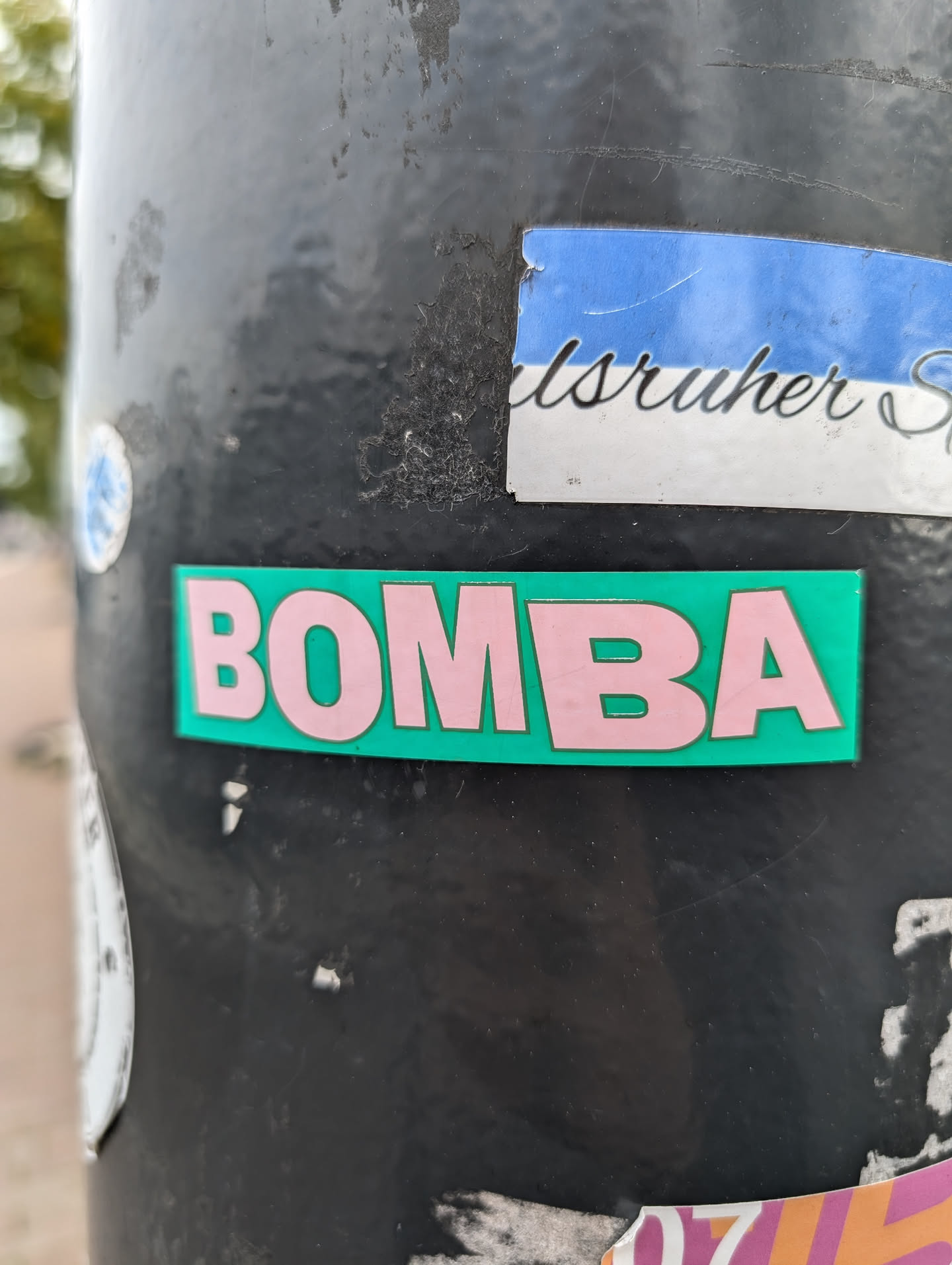 A teal and pink sticker with the word BOMBA in capital letters. The sticker is attached to a dark-colored pole.