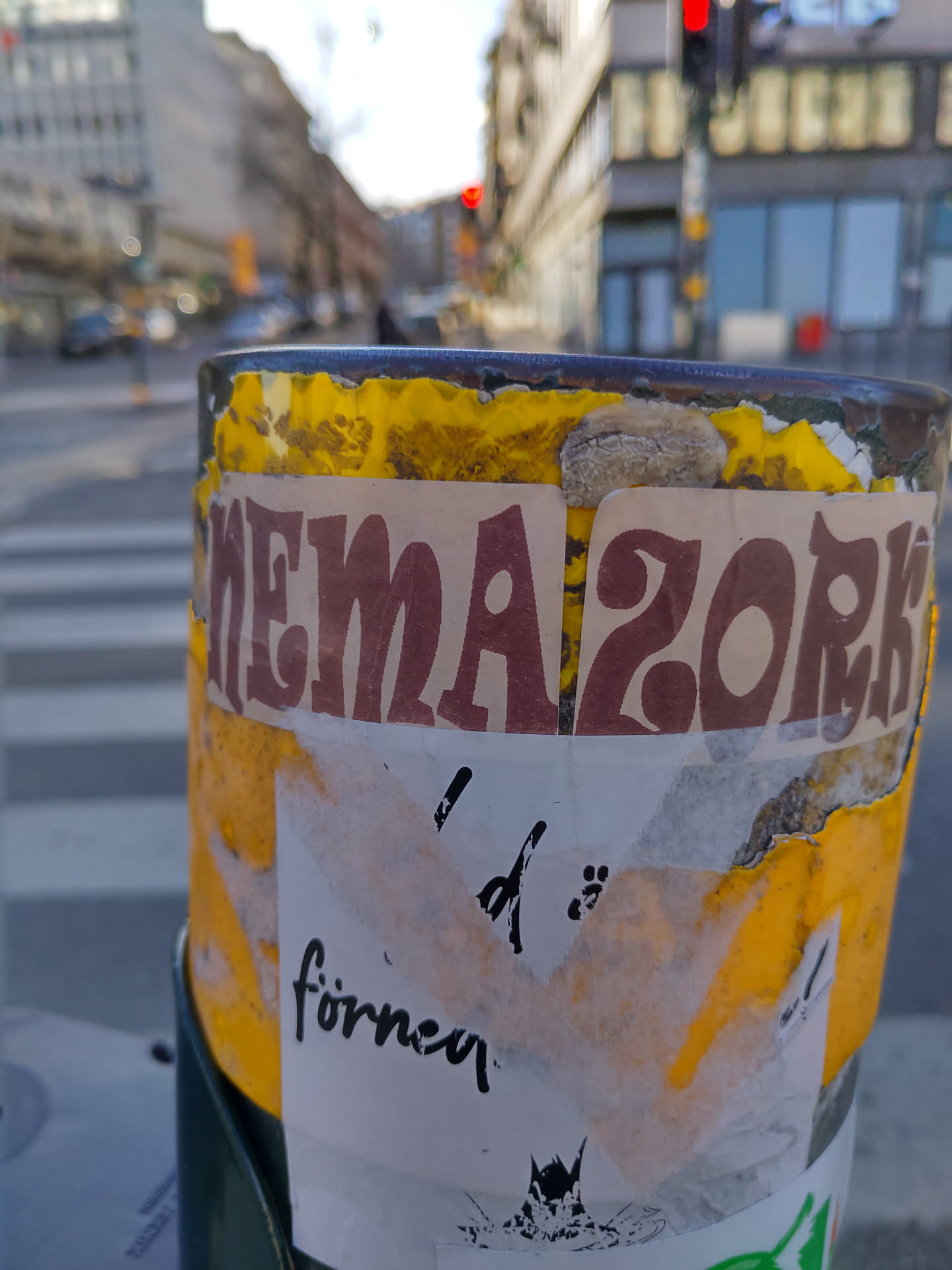 A close-up shot of a yellow pole with multiple stickers attached. The most prominent sticker features the word "Nemazork" in a bold, stylized font. A smaller, partially obscured sticker below it has some illegible text, but the word "förnen" is visible. The stickers appear weathered and slightly damaged, suggesting they have been exposed to the elements.