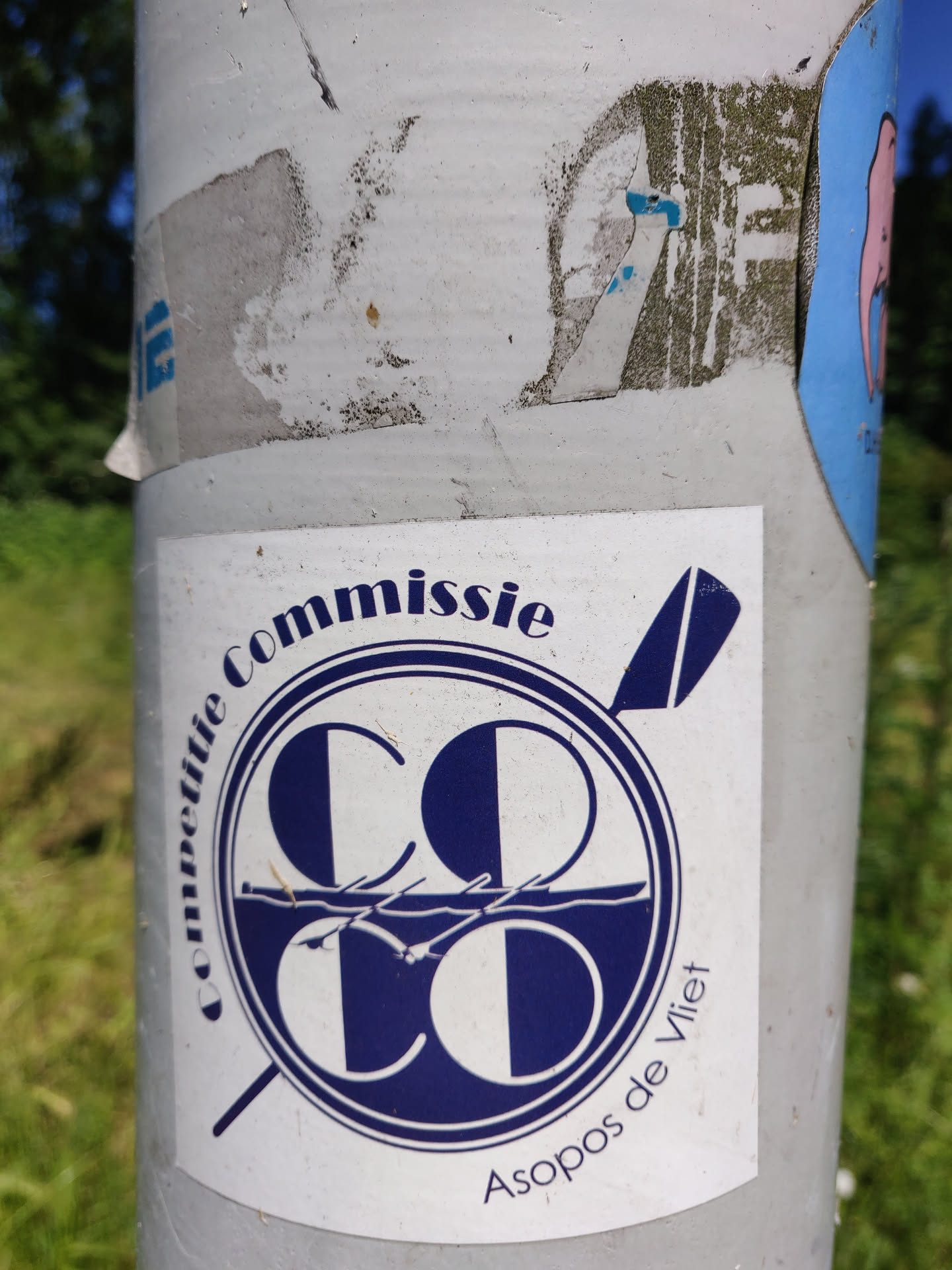 A sticker featuring the logo of Asopos de Vliet, a rowing club. The logo is a circular design with the letters 'CC' intertwined, with a depiction of rowing boats below.  The text 'Commissie' is written around the top of the circle and 'Asopos de Vliet' is written at the bottom.