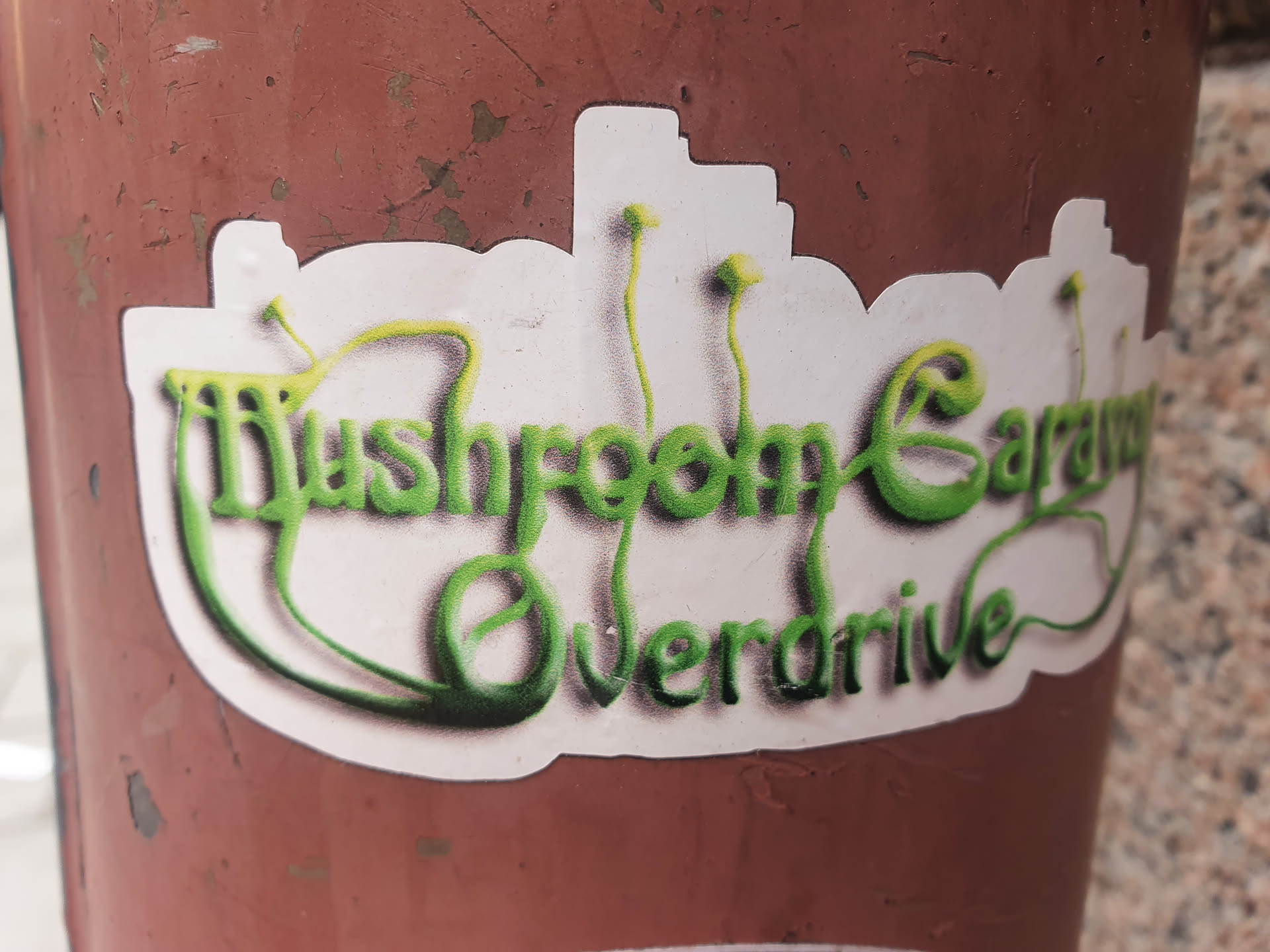 A sticker with the text "Mushroom Garage Overdrive" written in a stylized green font on a white background. The design includes small mushroom-like elements.