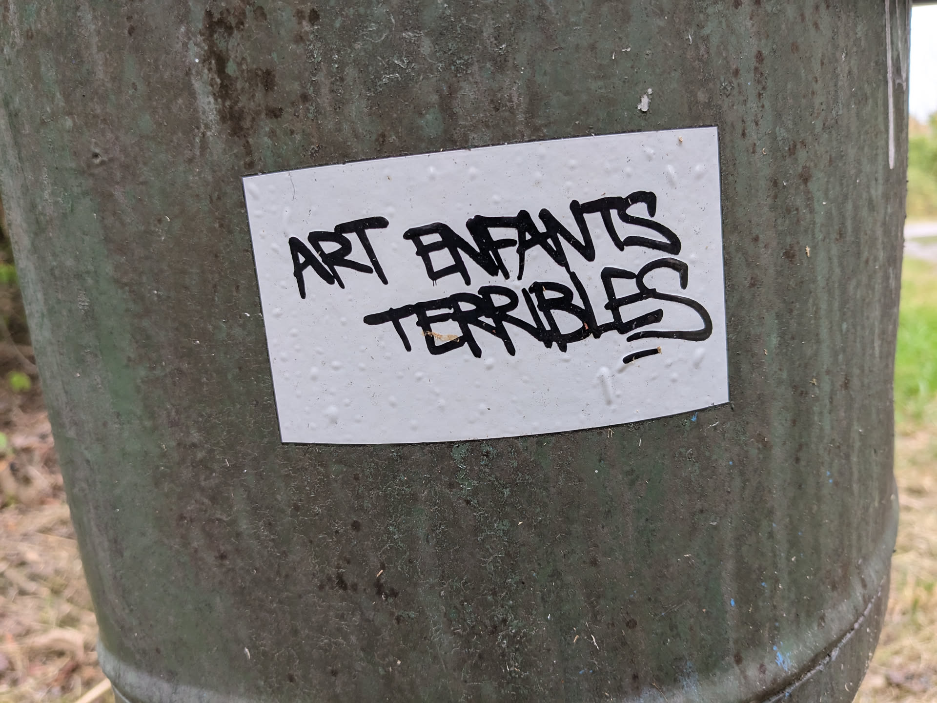 A white sticker with black graffiti-style text that says "Art Enfants Terribles". The sticker is affixed to a weathered, green metal surface.