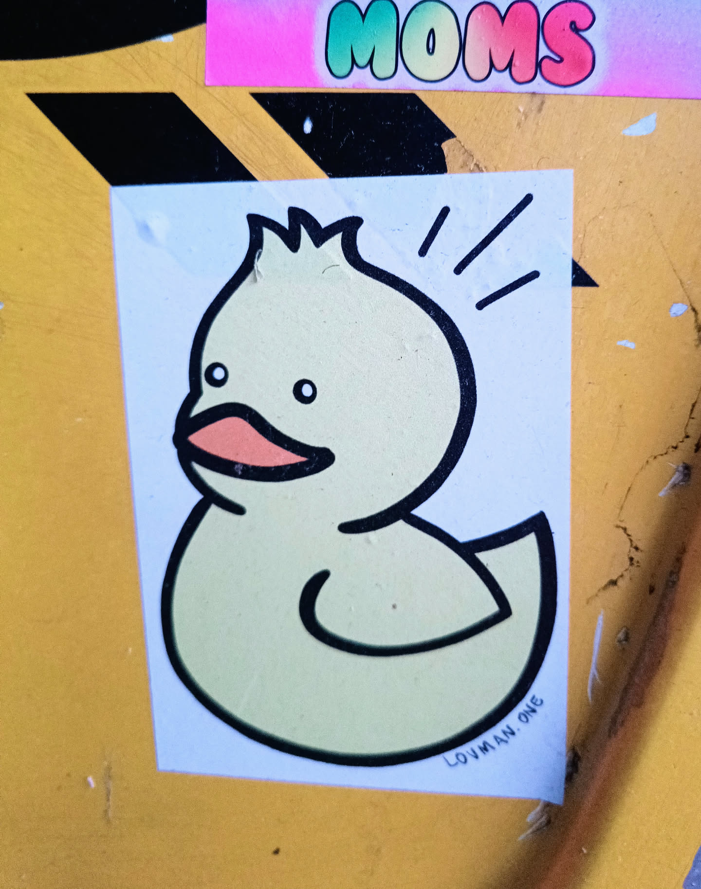 A simple sticker featuring a cartoon drawing of a cute yellow duck. The duck has a simplistic design with a few lines indicating movement or excitement. The artist's signature, LOVMAN.ONE, is visible at the bottom.