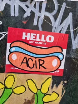 A sticker featuring a hot dog-shaped name tag with the name ACIR written on it. The sticker is red and white, and the hot dog is orange and light blue.