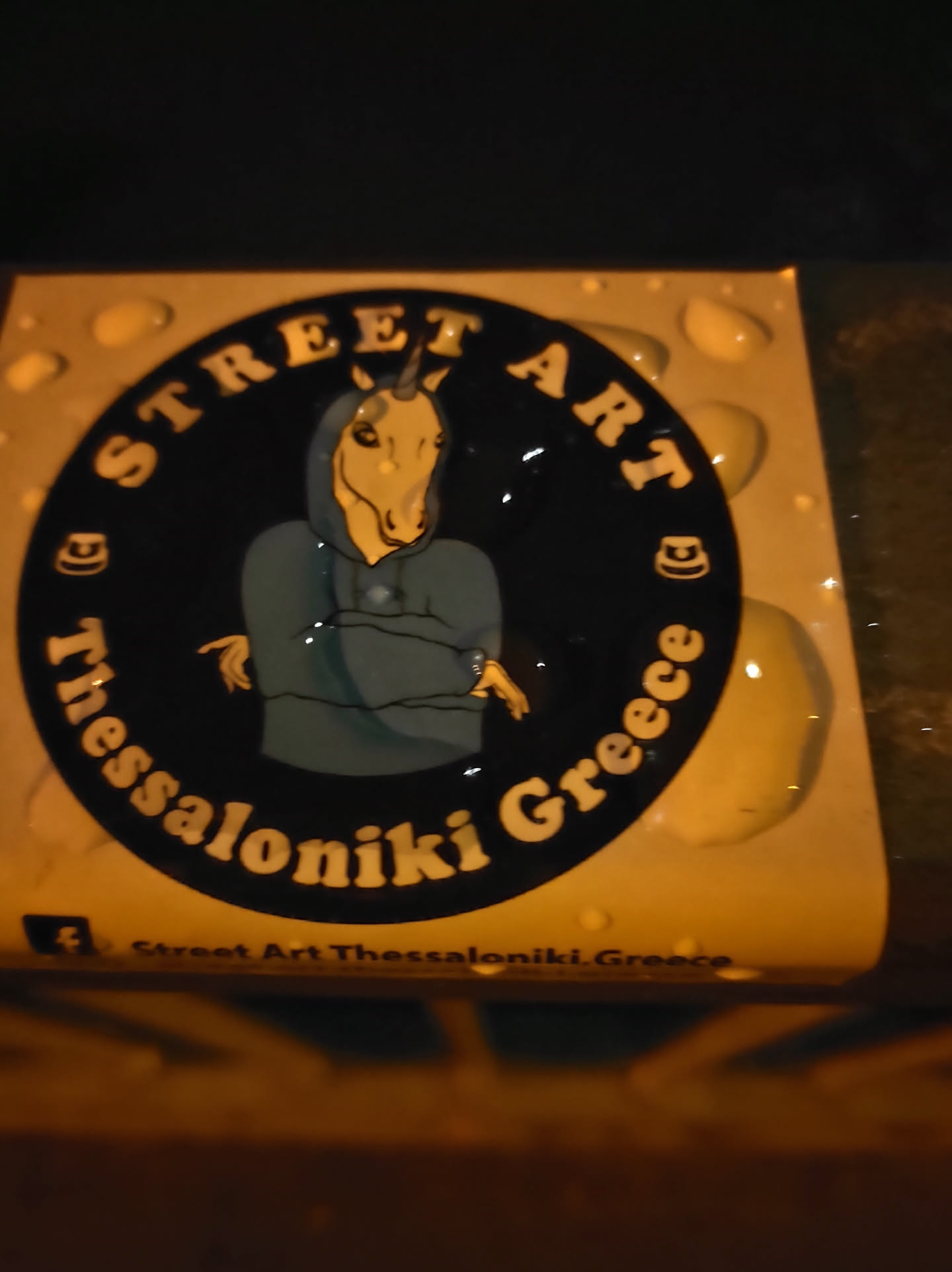 Round sticker featuring a unicorn wearing a hoodie. The sticker has a black background with white and yellow text.  The text says "Street Art" at the top, "Thessaloniki Greece" wrapped around the bottom, and has a small facebook logo at the bottom.
