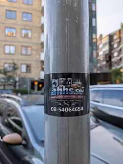 A black rectangular sticker wrapped around a metal pole. It features icons of household appliances and a hot tub at the top, followed by the blue website name 'ahhs.se'. Below, a red banner displays 'ÅKERSBERGA HUSHÅLLSSERVICE' above Swedish text detailing repair services, a crossed wrench and screwdriver graphic, and a large phone number at the bottom.