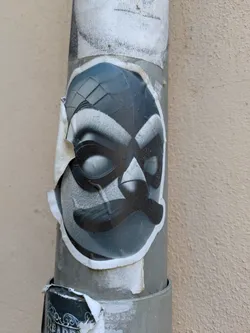 The sticker features a grayscale cartoon character with a mask and a mustache. The mask design includes diamond-shaped segments at the top and covers the eyes. The sticker is peeling off at the edges, and it's attached to a gray metal pipe that is also covered with other stickers.