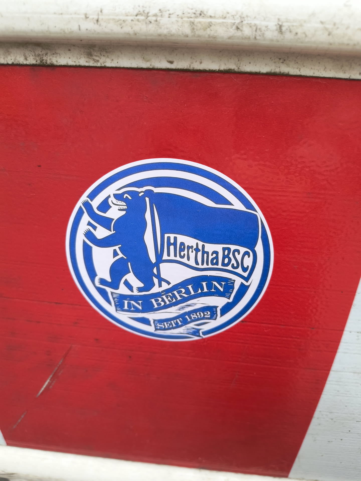 A blue and white circular sticker featuring the Hertha BSC logo, which includes a bear and a flag.  The text 'HerthaBSC', 'IN BERLIN', and 'SEIT 1892' is visible. The sticker is affixed to a red and white surface.