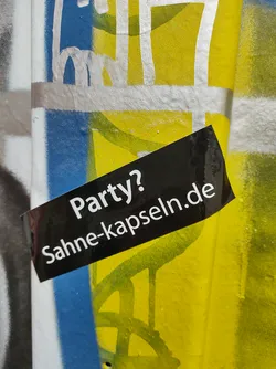 A black sticker on a graffitied wall. The sticker has white text that says "Party? Sahne-kapseln.de".