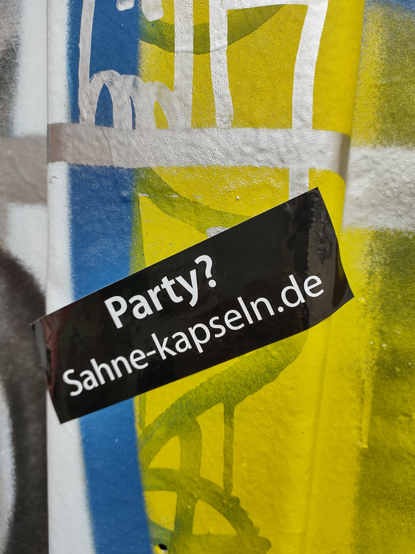 A black sticker on a graffitied wall. The sticker has white text that says "Party? Sahne-kapseln.de".