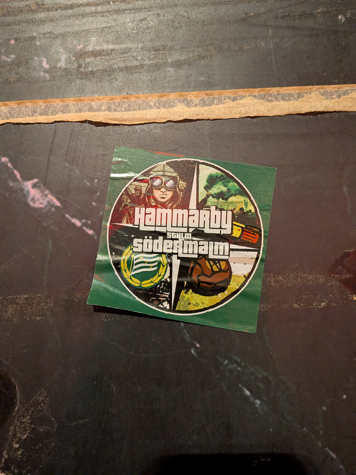 The sticker is circular with a green background. The top section features a stylized illustration of a pilot wearing goggles and a helmet, with other figures in the background. Below the pilot, the text 'HAMMARBY' is prominently displayed in a bold, white, blocky font reminiscent of the Grand Theft Auto logo. Beneath this, 'SÖDERMALM' is written in a similar style. The bottom left quadrant contains a circular emblem with green and white stripes, surrounded by a yellow laurel wreath. The bottom right quadrant depicts a vintage-style football with a brown and white pattern, set against a yellow and green background with abstract black elements.