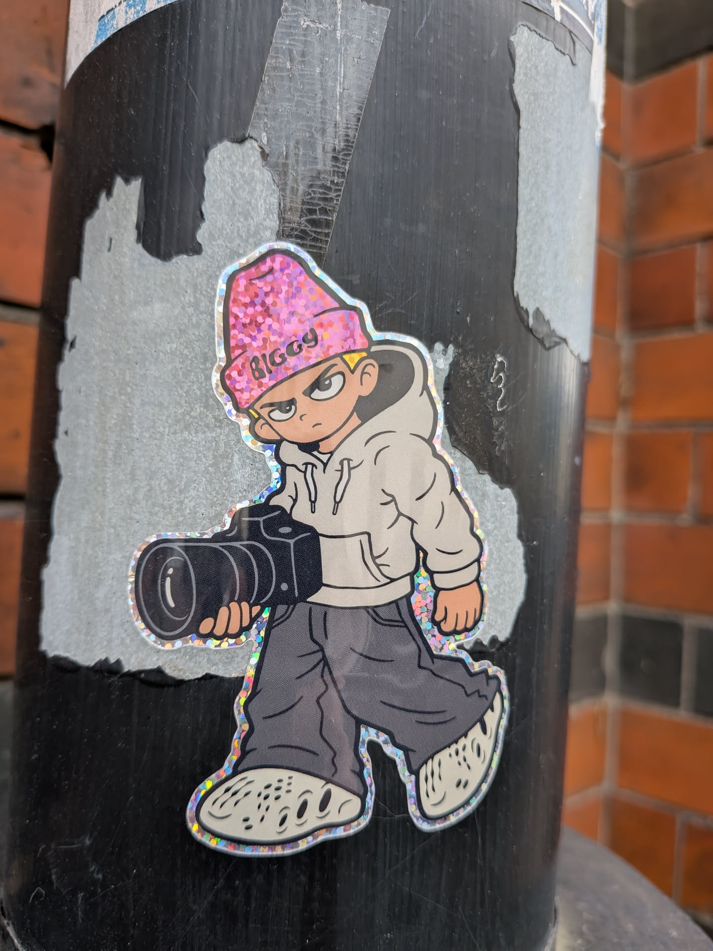The sticker features a cartoon-style illustration of a young person wearing a pink, glittery beanie with the word 'BIGGY' in black lettering. They have a determined expression with furrowed brows and sharp eyes. The character is dressed in a light-colored hoodie, dark gray baggy pants, and distinctive white clogs with holes. They are holding a large black camera with a prominent lens in their right hand. The sticker has a holographic border that shimmers with rainbow colors, giving it a distinct, eye-catching appearance.