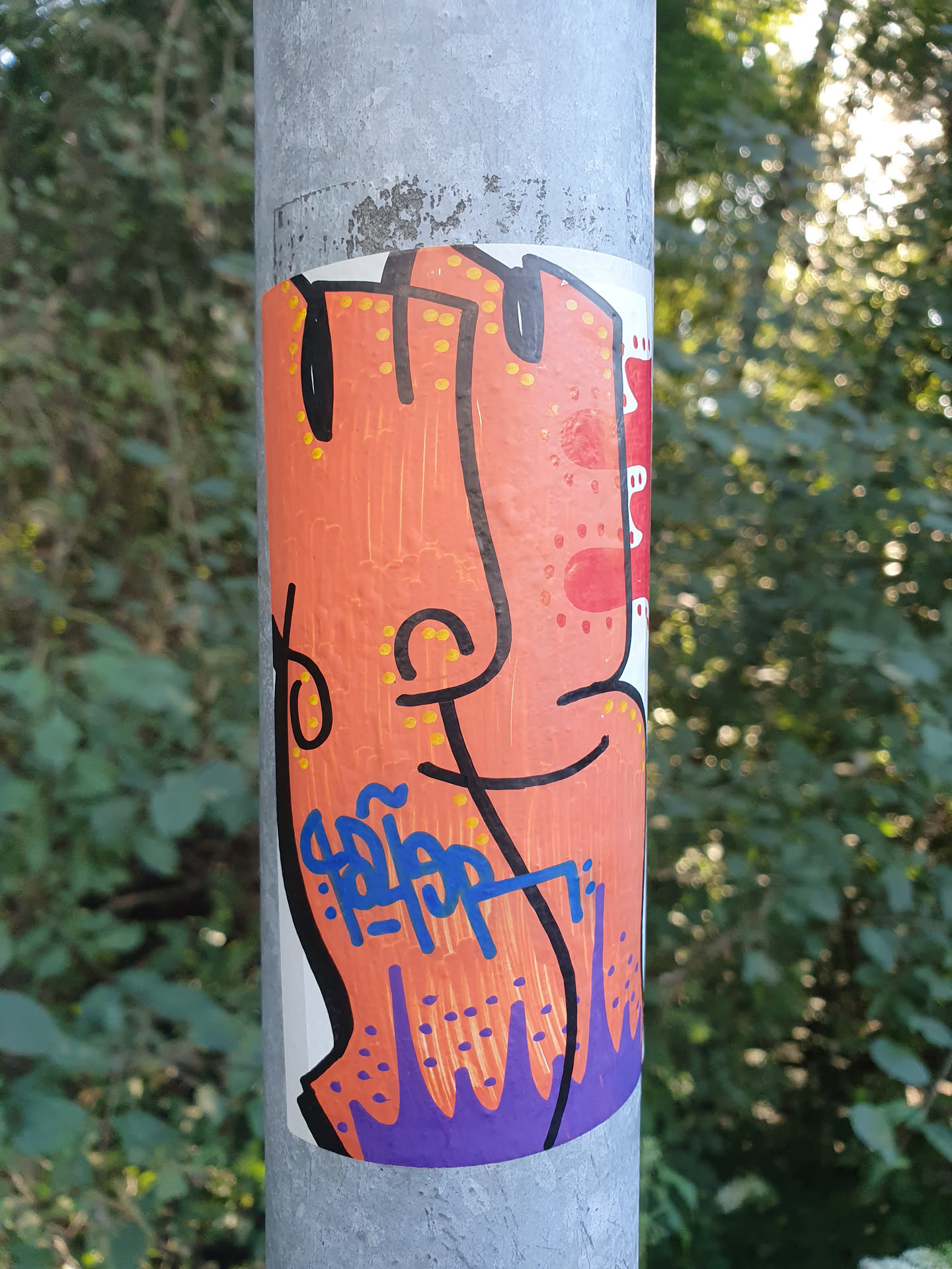A sticker featuring an abstract orange figure with a purple bottom, reminiscent of dripping paint, is affixed to a gray metal pole.  The figure has a simplified face and body with small yellow dots scattered across its surface. The artist's signature, which appears to read 'Pippop', is written in blue ink on the sticker. The background shows green foliage.