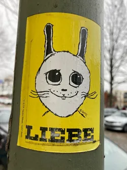 A yellow sticker with a cartoon bunny and the word "Liebe" (German for Love) in bold black letters. The bunny has big eyes and a simple smile. The artist's signature is visible in small writing at the bottom right. The sticker is applied to a gray pole, possibly on a street in Berlin.