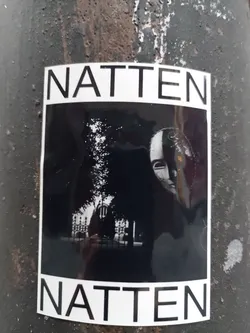 Black and white photo sticker with the word "NATTEN" at the top and bottom. The photo depicts a person's face partially obscured by shadows, with a building and trees visible in the background. 
