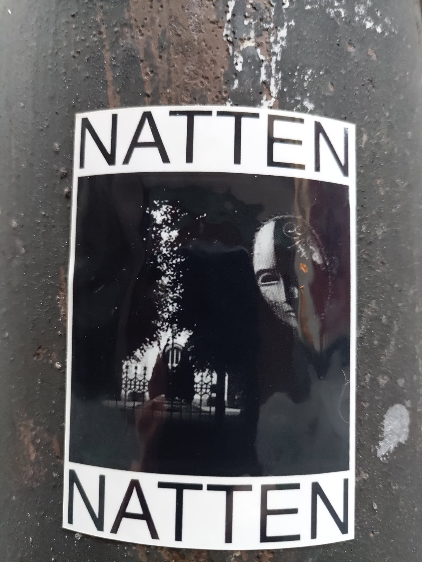 Black and white photo sticker with the word "NATTEN" at the top and bottom. The photo depicts a person's face partially obscured by shadows, with a building and trees visible in the background. 