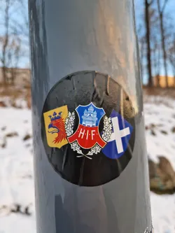 A round sticker affixed to a gray pole. The sticker features a central emblem with the letters "H.I.F." surrounded by wheat stalks, flanked by what appear to be regional or historical crests. The colors are primarily black, red, blue, and yellow. The sticker shows signs of wear and tear.