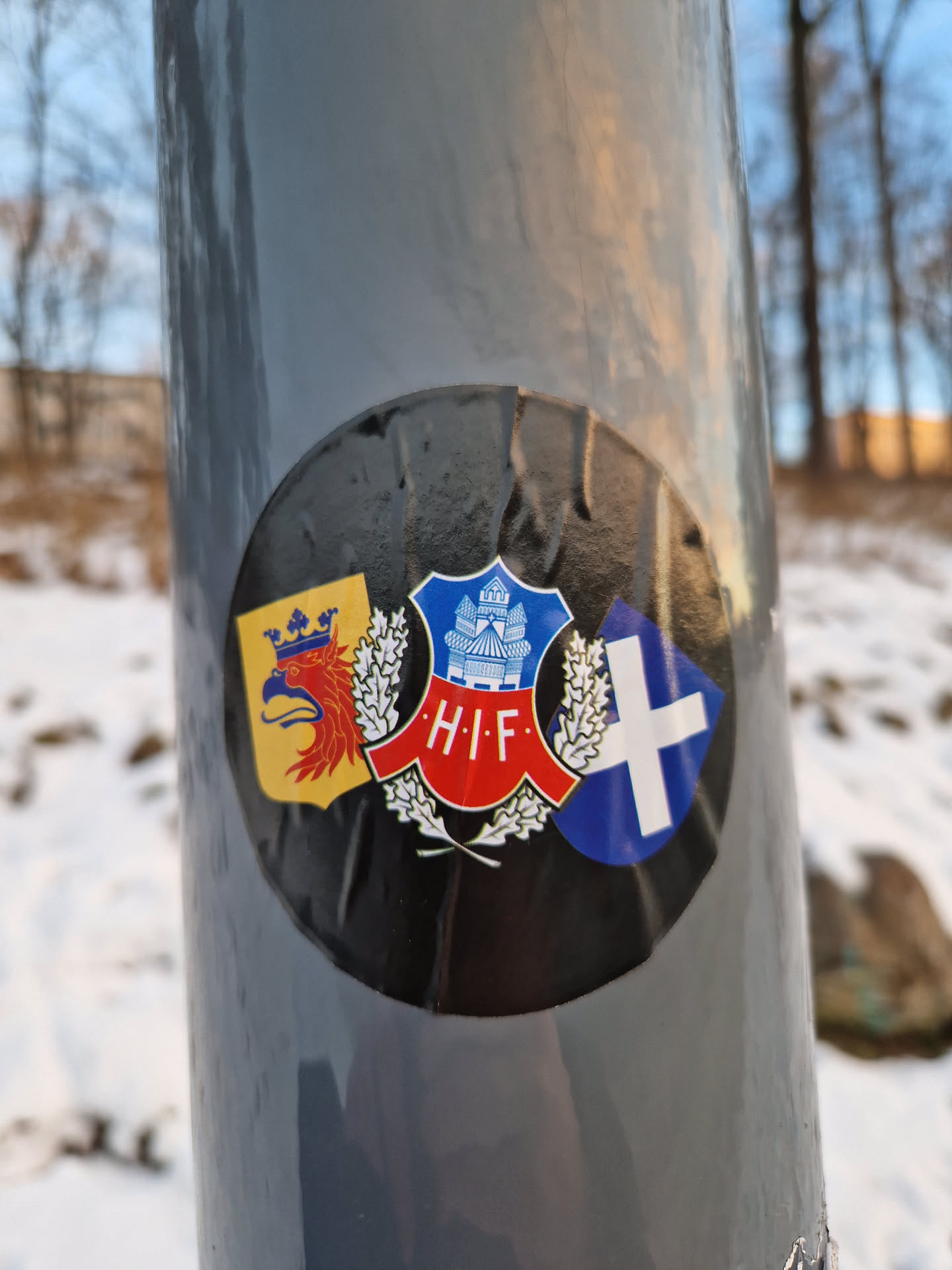 A round sticker affixed to a gray pole. The sticker features a central emblem with the letters "H.I.F." surrounded by wheat stalks, flanked by what appear to be regional or historical crests. The colors are primarily black, red, blue, and yellow. The sticker shows signs of wear and tear.