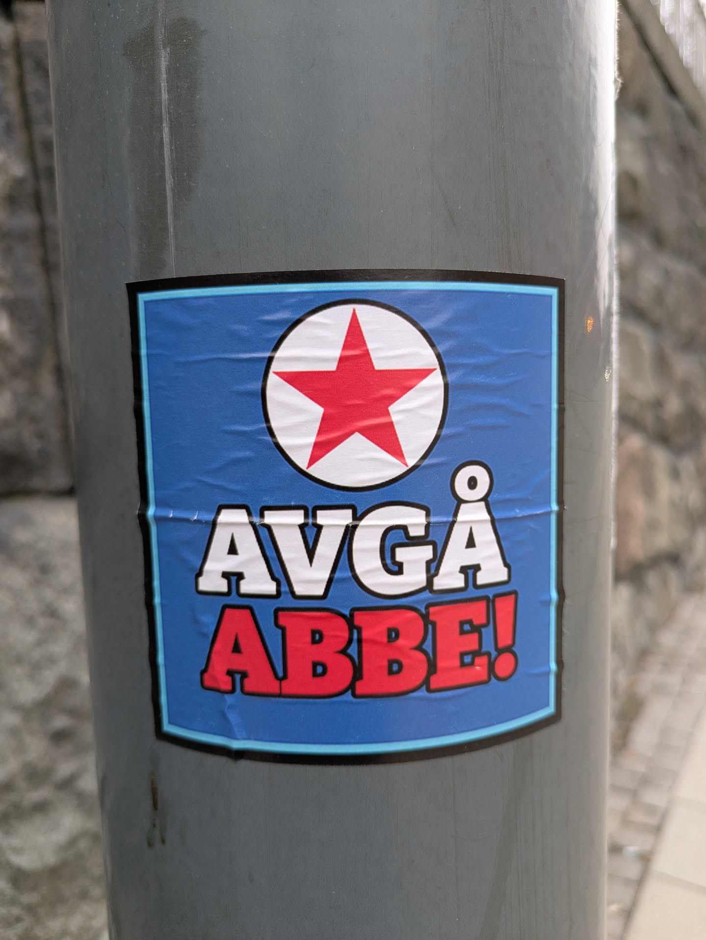 A sticker with a blue background and a white circle containing a red star at the top. Below the star, the text "Avgå" is written in white, and "Abbe!" is written in red. The sticker is affixed to a gray pole.