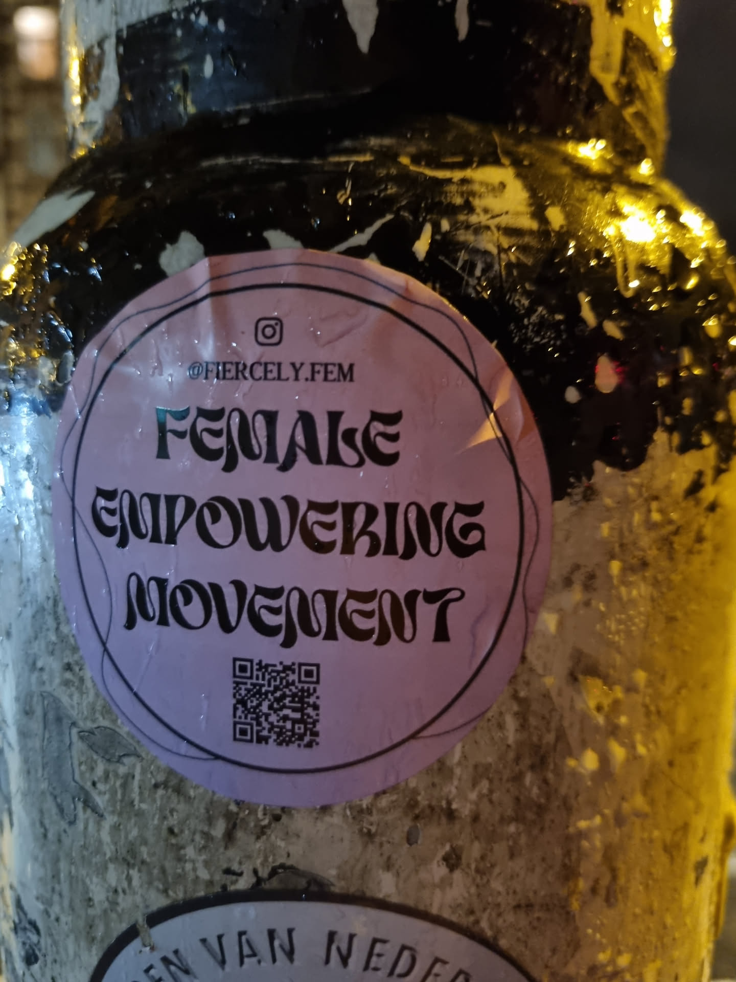 A round pink sticker with the text "FEMALE EMPOWERING MOVEMENT" in a stylized font. It also includes an Instagram handle (@fiercely.fem) and a QR code. The sticker is affixed to a weathered surface.