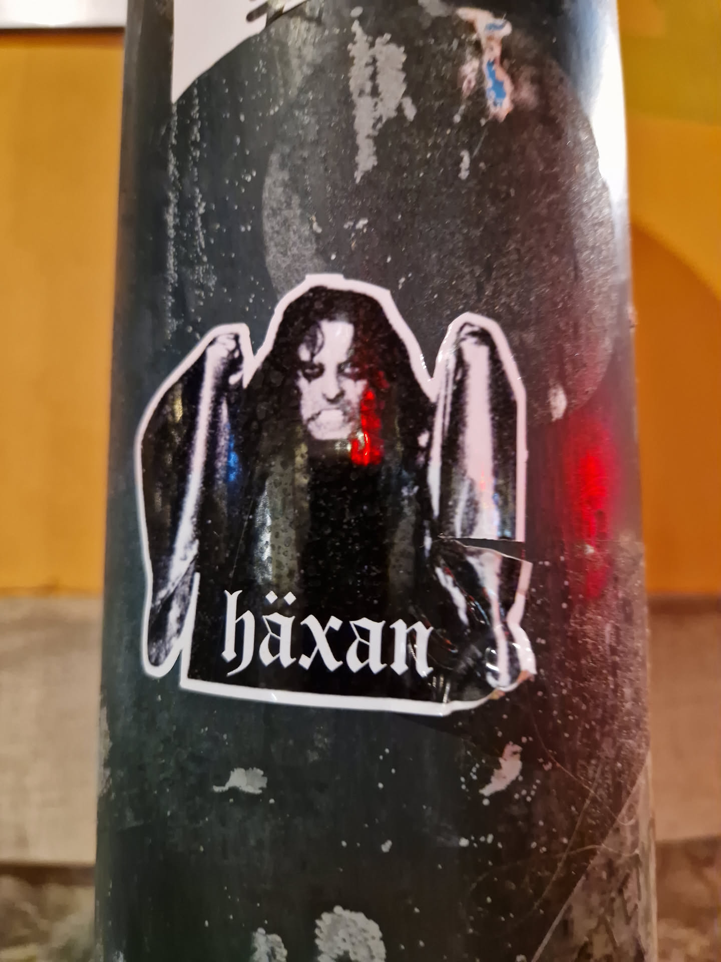 A black and white sticker featuring a figure reminiscent of a witch or dark entity, with the word "Häxan" prominently displayed at the bottom. The sticker is affixed to a dark-colored surface, possibly a pole or post.  The image quality is slightly affected by the surface texture and some minor damage to the sticker itself.