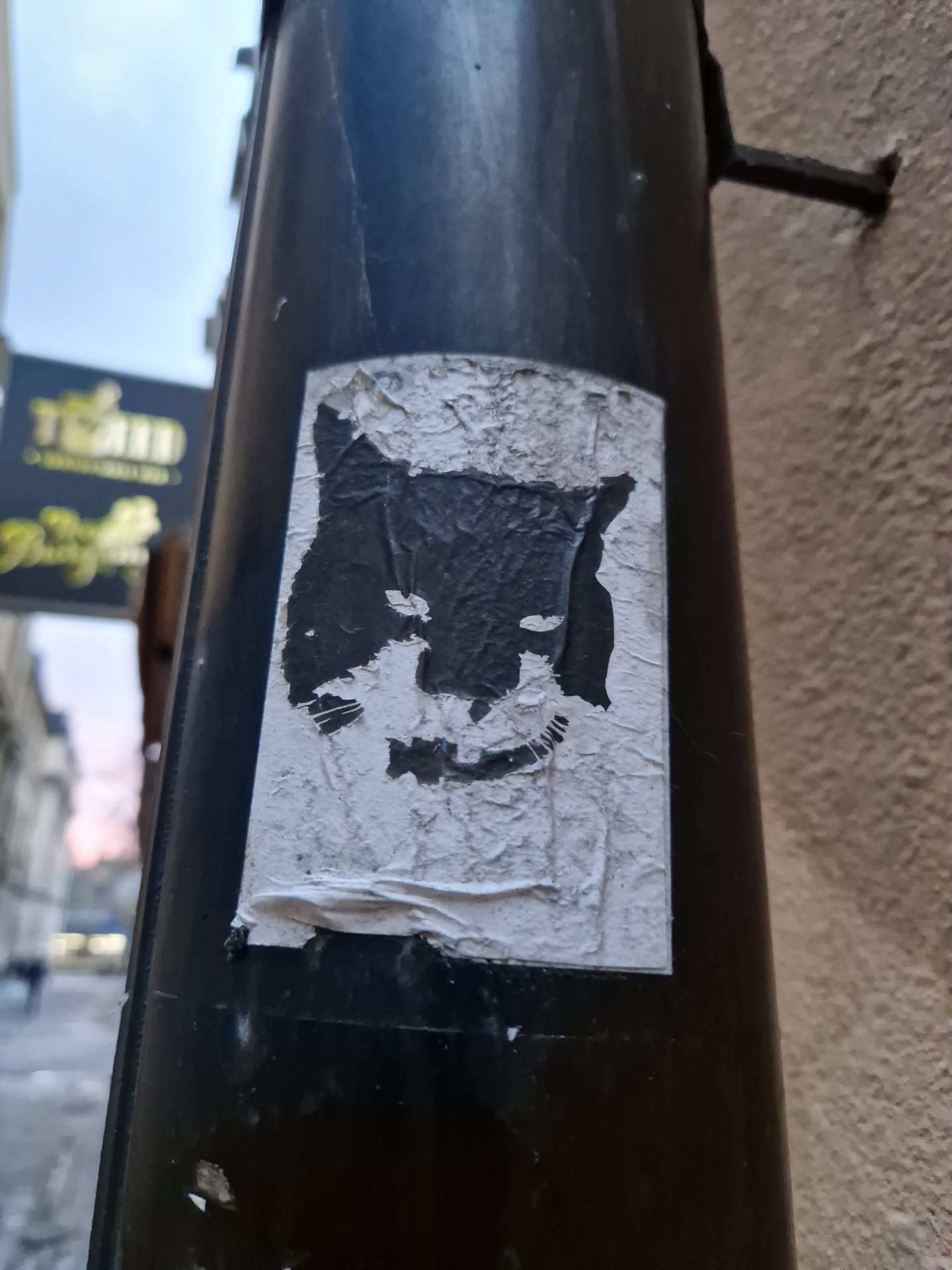 A partially peeled, weathered sticker of a wolf's head in black and white. The sticker is adhered to a dark-colored pole, and some of the edges are peeling off, revealing the surface underneath. The wolf's face is stylized and has a slightly menacing appearance.