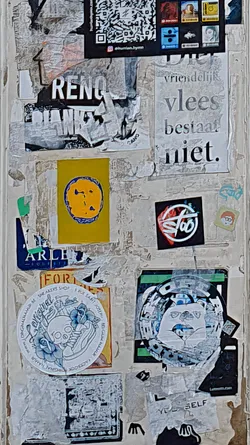 A cluttered wall covered with various stickers and torn posters. The stickers feature diverse designs, including a skull with flowers, abstract patterns, and text in different fonts. The overall aesthetic is chaotic and urban, reflecting the nature of street art.
