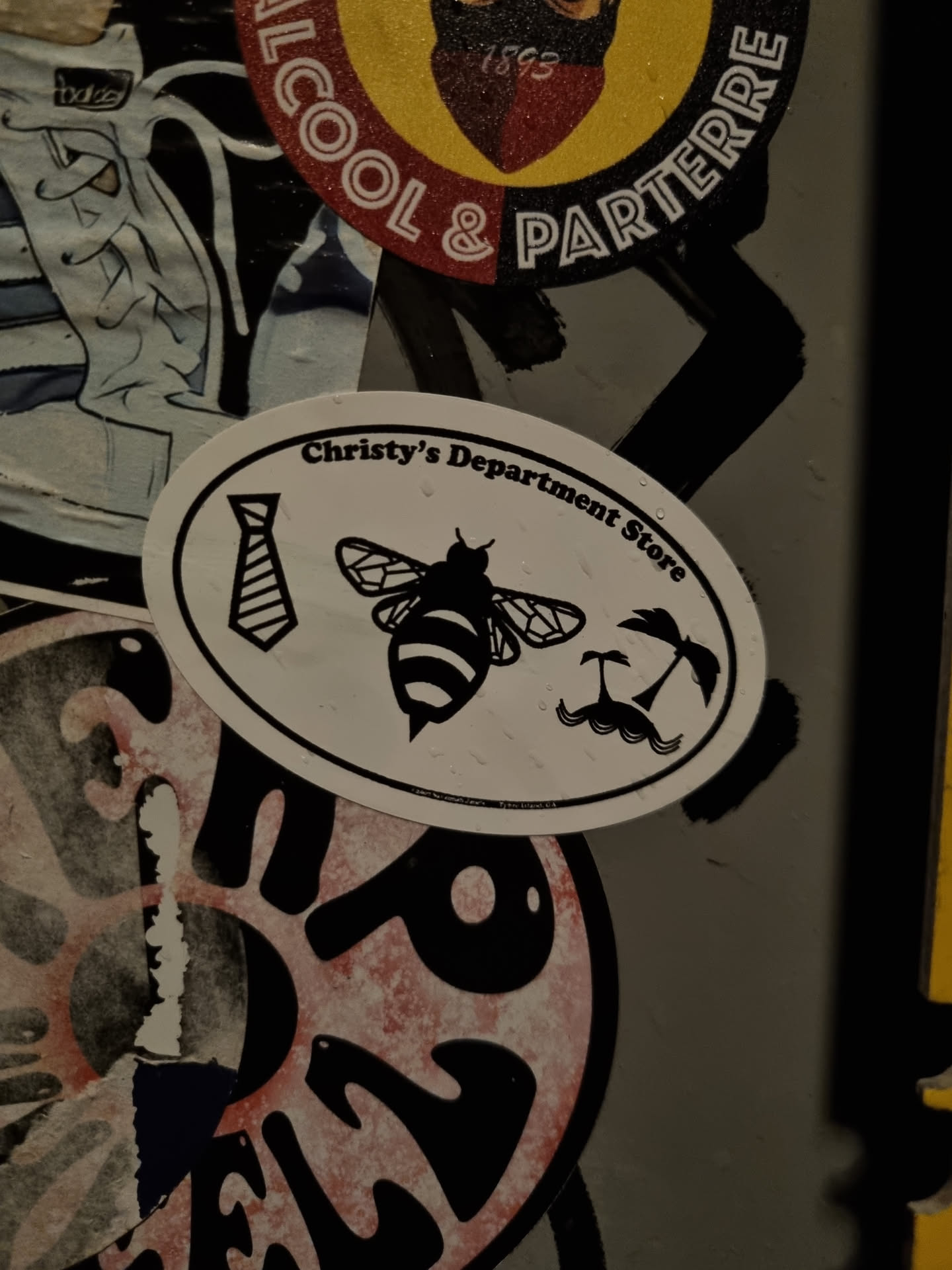 Oval sticker featuring a tie, a bee, and palm trees.  The sticker is white with black illustrations. 
