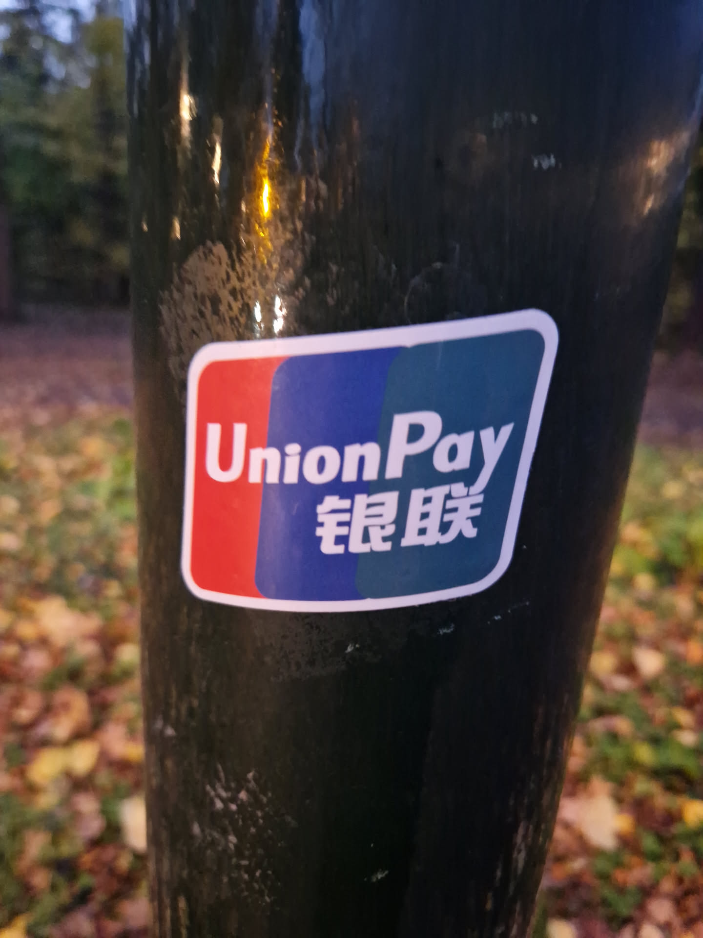 A rectangular sticker with the UnionPay logo. The logo is mainly blue and red, with the words "UnionPay" in English and the Chinese characters for "银联" (Yínlián) below it. The sticker is affixed to a dark-colored pole outdoors.