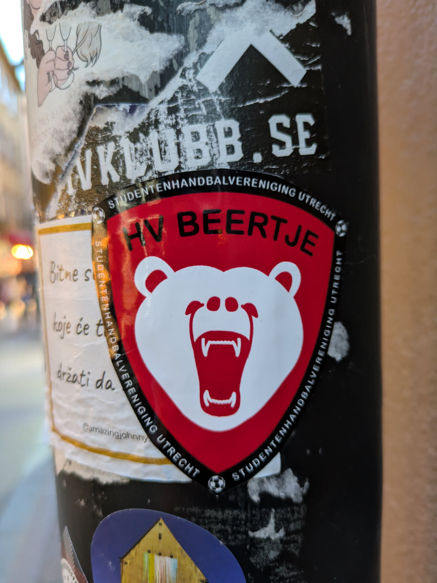 The central sticker features a shield emblem with a fierce white bear's head as its primary graphic. The bear has its mouth wide open, revealing sharp teeth and a red interior. Its eyes are black dots, and it has two small ear-like protrusions. The shield itself is outlined in black, with a red background that fades to white around the bear's head. Along the upper edge of the shield, in black lettering on a white curved banner, is text. Similar black text also curves along the bottom edge of the shield and up the sides, interrupted by small black and white soccer ball graphics on the left and right sides. The overall impression is that of a sports team logo.