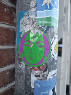 A round sticker featuring a stylized face with closed eyes and a subtle smile. The face is lime green with purple hair, creating a striking contrast. The sticker is adhered to a weathered metal pole, surrounded by other stickers and graffiti, adding to its urban appeal.