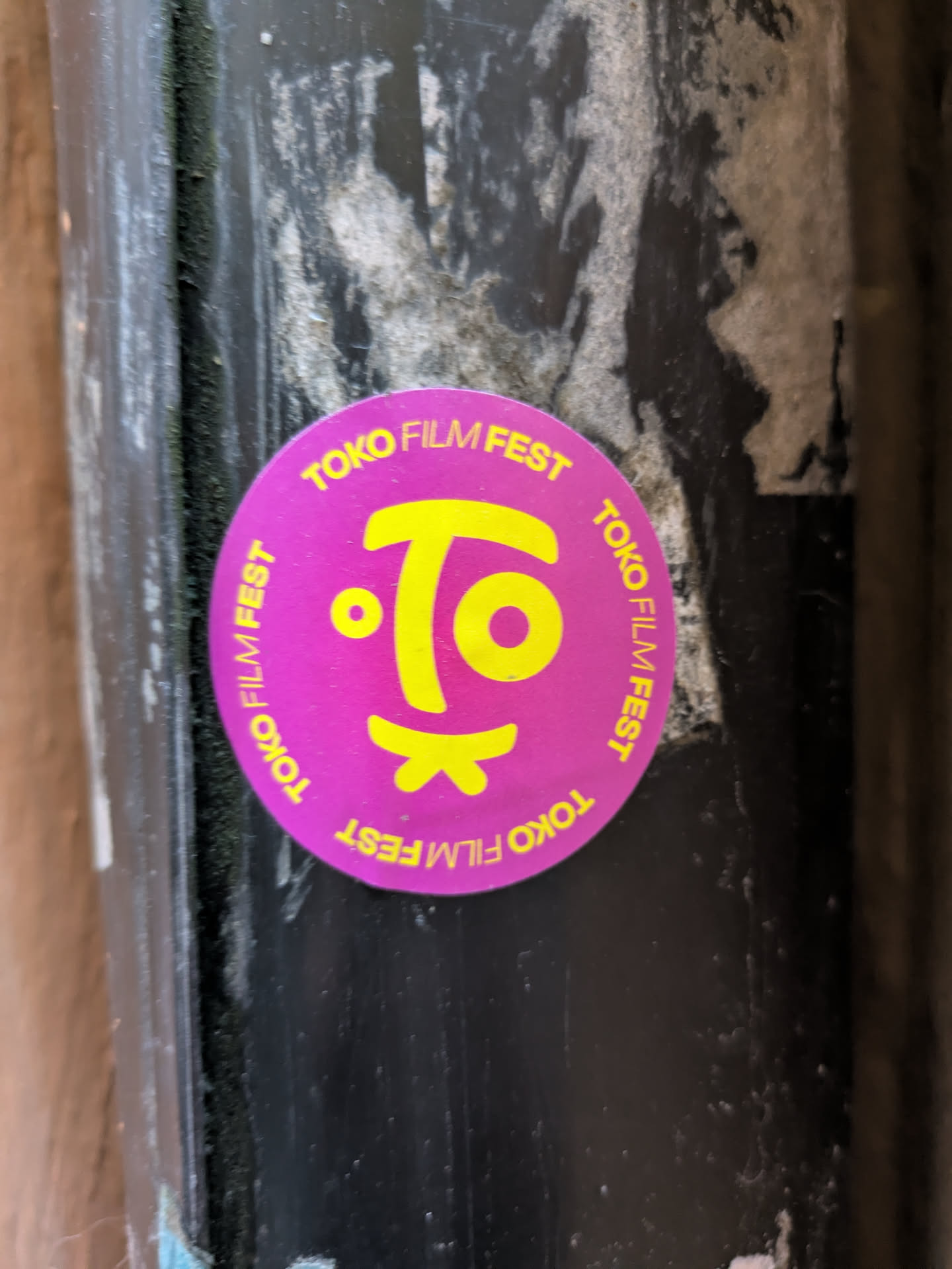 A round sticker with the text "Tokyo Film Fest" repeated around the perimeter and a stylized logo in the center. The sticker is purple with yellow text and logo. It's affixed to a dark-colored surface.