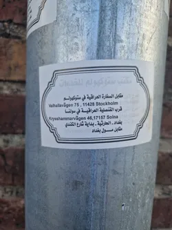 A sticker on a metal pole provides addresses in both Arabic and English for an office located near the Iraqi embassy and consulate in Stockholm and Solna, Sweden.  Phone number is also included.
