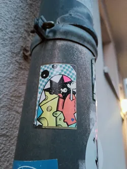 A sticker featuring a cartoon monster with big teeth, surrounded by cheese pieces. The style is colorful and playful, with a mix of geometric and organic shapes. The sticker is applied to a metal pipe outdoors.