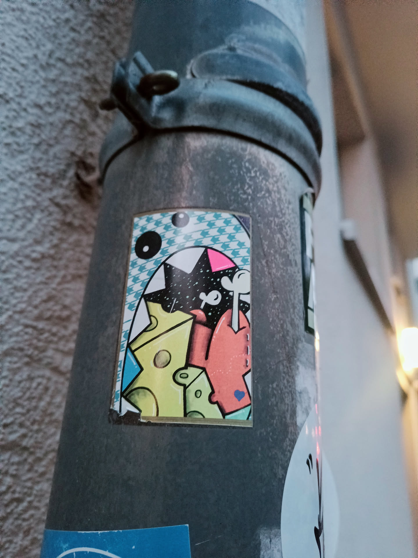 A sticker featuring a cartoon monster with big teeth, surrounded by cheese pieces. The style is colorful and playful, with a mix of geometric and organic shapes. The sticker is applied to a metal pipe outdoors.