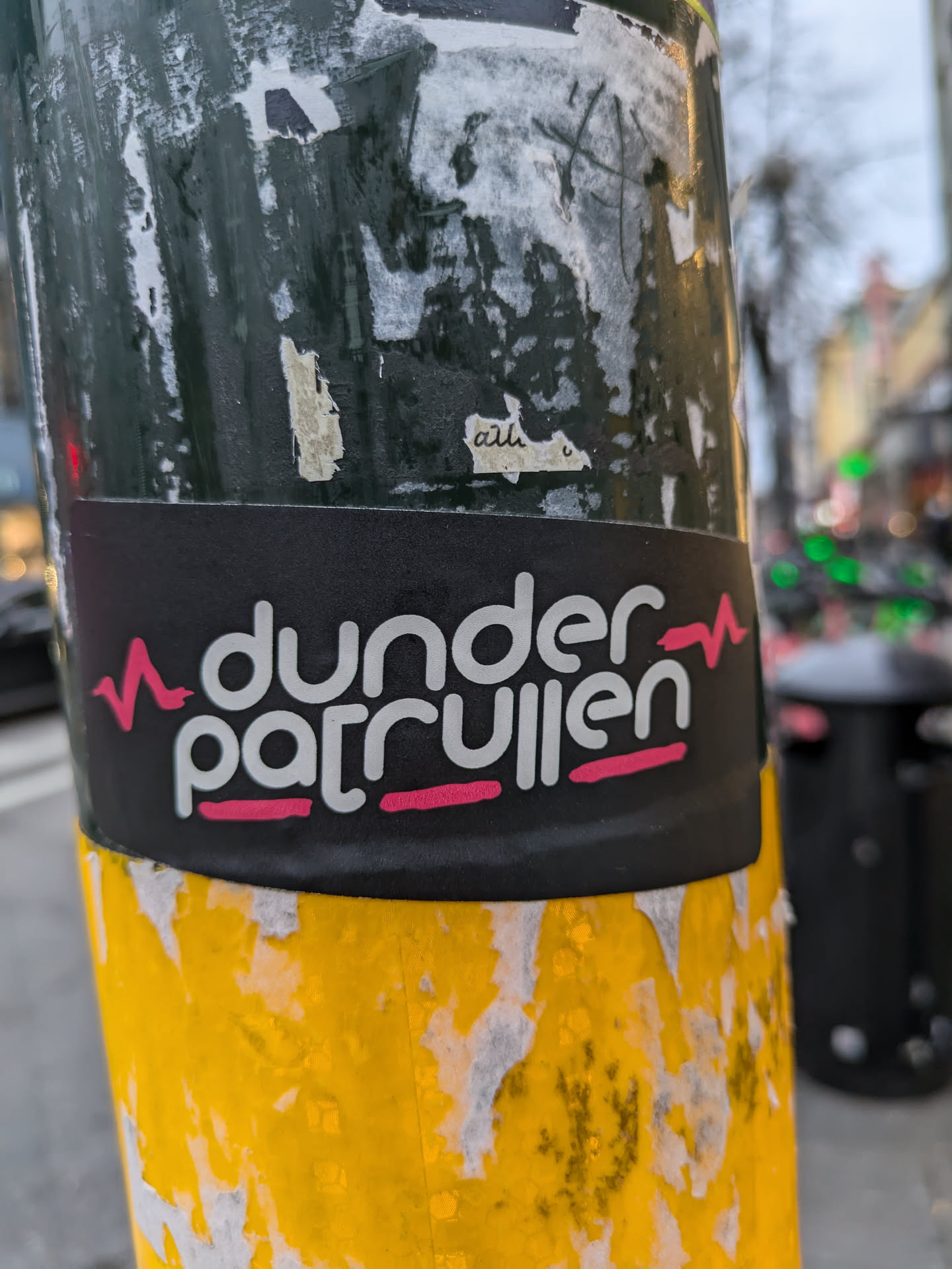 A black and pink sticker with the text "dunder patrullen" and a heartbeat symbol on each side. The sticker is applied to a yellow pole.