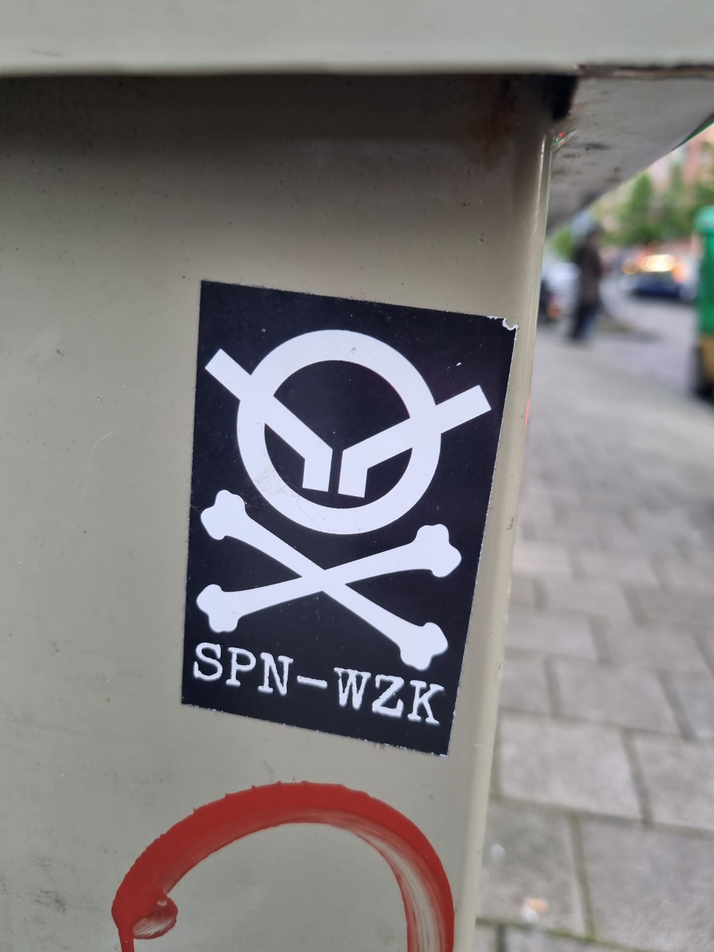 A black and white sticker with a stylized skull and crossbones graphic. The text 'SPN-WZK' is printed below the graphic. The sticker is affixed to a light beige surface outdoors.