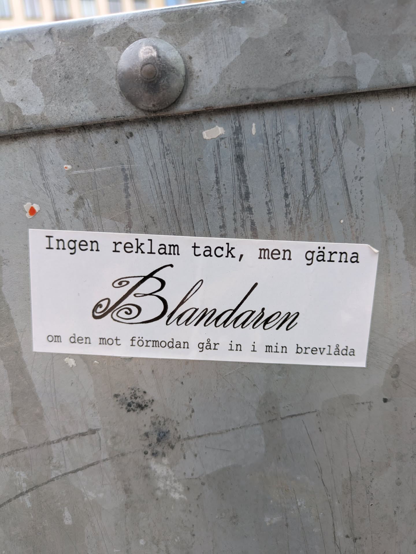 A white rectangular sticker is affixed to a gray metal surface. The sticker has black text in a serif font. The text reads "Ingen reklam tack, men gärna Blandaren om den mot förmodan går in i min brevlåda".