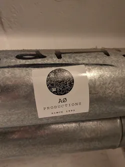 Street sticker A square white sticker adhered to a galvanized metal pipe. The sticker features a circular black-and-white QR code centered at the top. Below the circular code, black serif text reads 'AØ' above 'PRODUCTIONS', followed by 'SINCE 1992' in smaller capital letters.