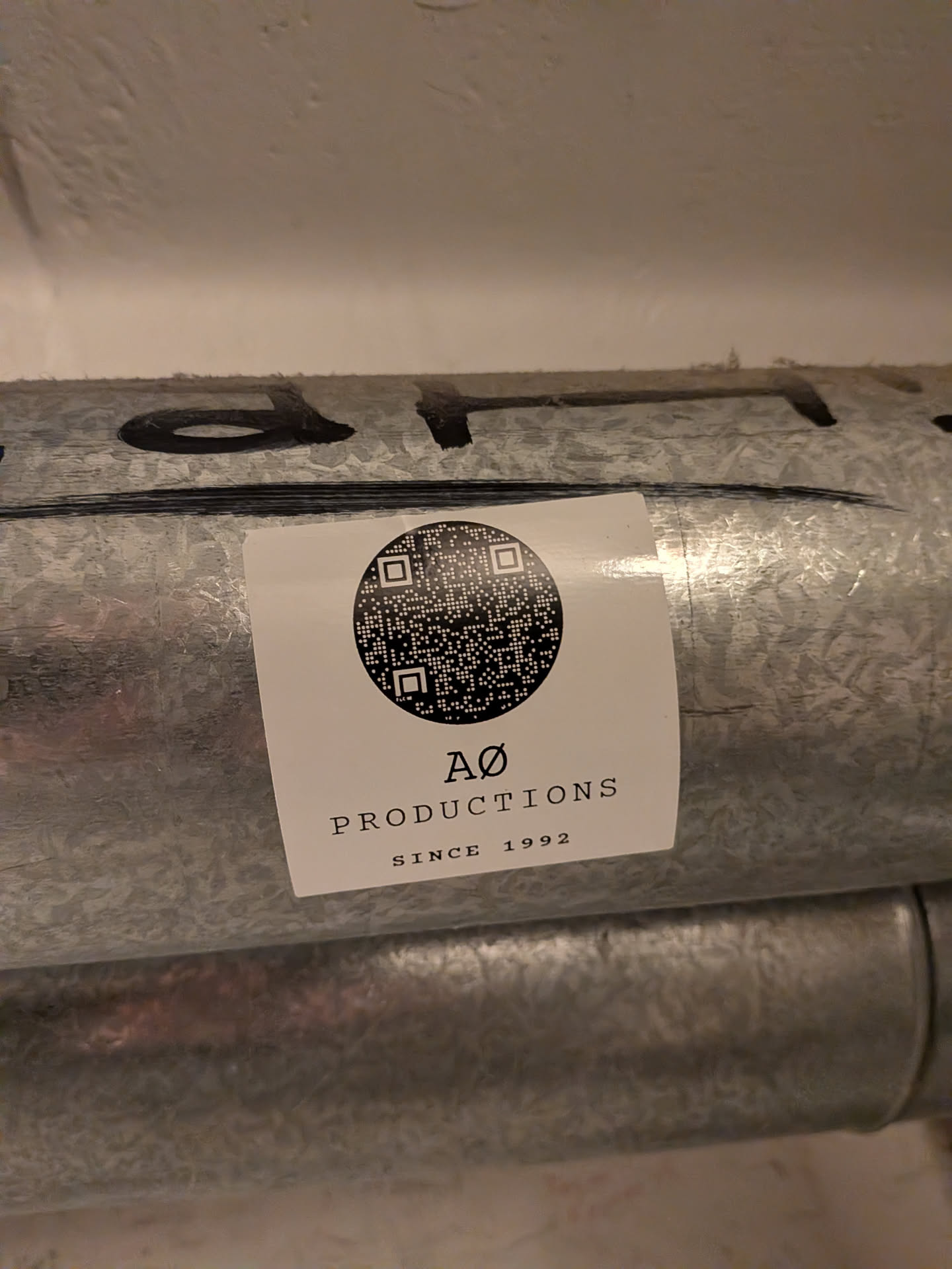 A square white sticker adhered to a galvanized metal pipe. The sticker features a circular black-and-white QR code centered at the top. Below the circular code, black serif text reads 'AØ' above 'PRODUCTIONS', followed by 'SINCE 1992' in smaller capital letters.