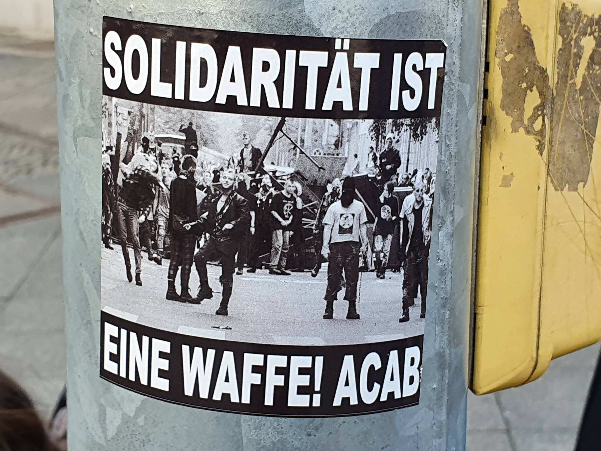 Black and white photo of a protest or riot with text overlay. The text on the sticker translates to: 'Solidarity is a weapon! ACAB'